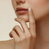 Gros plan sur une main féminine avec un anneau en or orné de diamants, portant un vernis à ongles nude. La femme touche délicatement son visage, mettant en valeur cet élégant bijou. Cette image évoque la beauté et le glamour associés aux accessoires de mode, idéale pour les collections de bijoux haut de gamme.