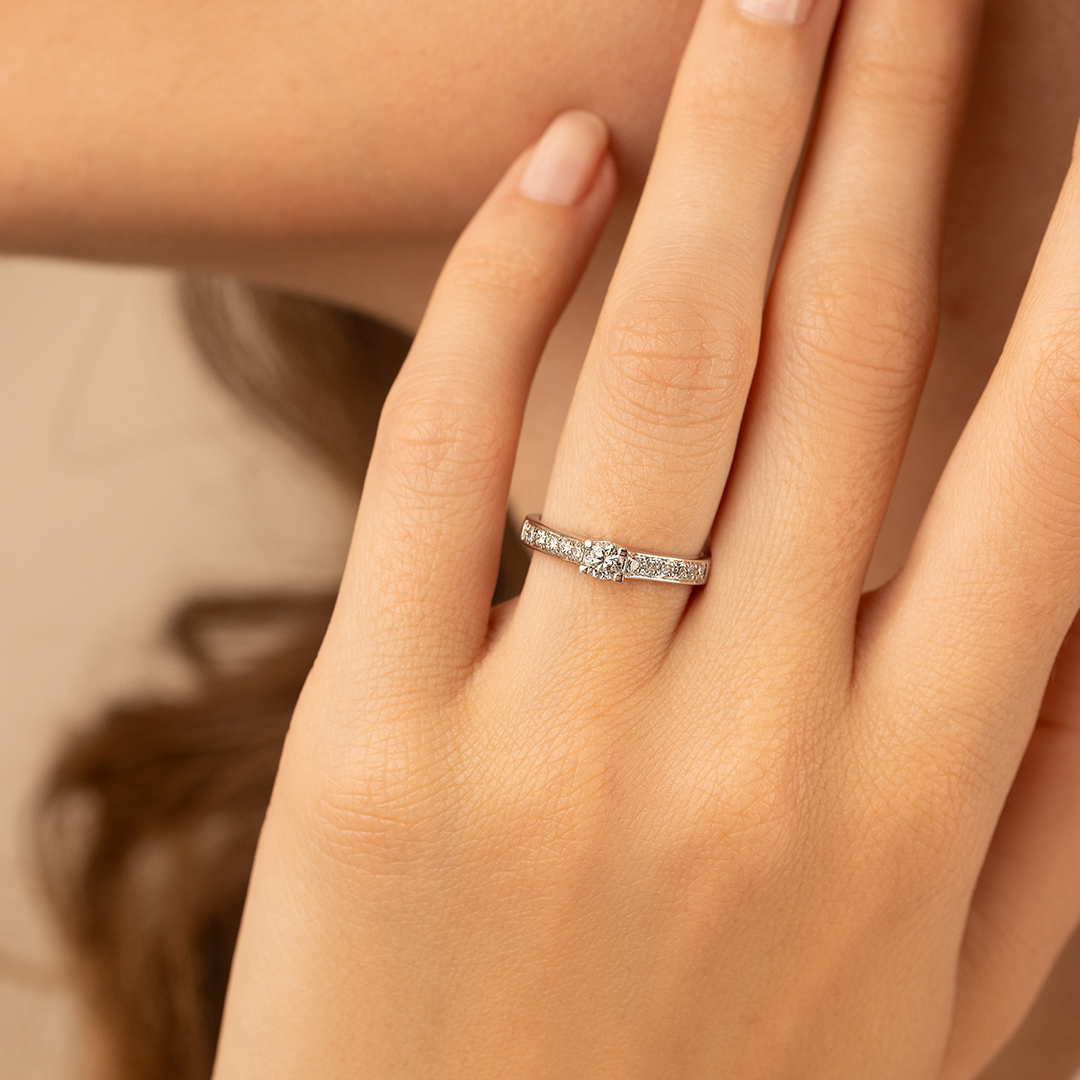 Bague en diamant élégante portée sur la main d'une personne, mettant en avant un zircon central scintillant entouré de petites pierres précieuses, modèle contemporain avec un design délicat, idéal comme bague de fiançailles ou bijou de luxe.