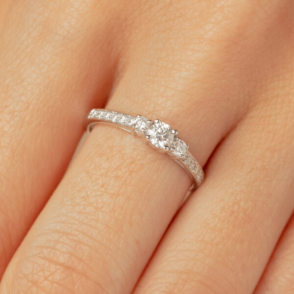 Bague en or blanc ornée d'un diamant central rond, flanquée de petites pierres scintillantes, portée sur un doigt. L'élégance et la brillance des diamants confèrent à cette bague un style classique et raffiné, idéale pour les fiançailles ou les occasions spéciales. Modèle de bijou contemporain avec une finition polie et des détails délicats.