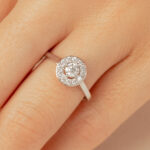 Bague en diamant avec un design élégant, présentant une pierre centrale entourée de petits diamants, sur un fond de main. Parfait pour les fiançailles ou une occasion spéciale, ce modèle classique en or blanc ou argent met en valeur la brillance des diamants tout en offrant une allure sophistiquée.
