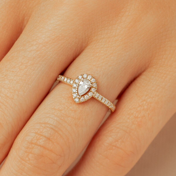 Bague en or avec design en forme de poire, sertie d'un diamant central entouré de petites pierres brillantes, portée sur une main. Idéale pour des occasions spéciales comme des fiançailles ou des anniversaires.