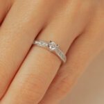 Bague en diamant élégante avec un design fin, mettant en valeur un diamant brillant au centre et des petits diamants sur la bande, positionnée sur un doigt humain. Parfaite pour une demande en mariage ou une occasion spéciale, ce modèle diamant reflecte sophistication et amour.