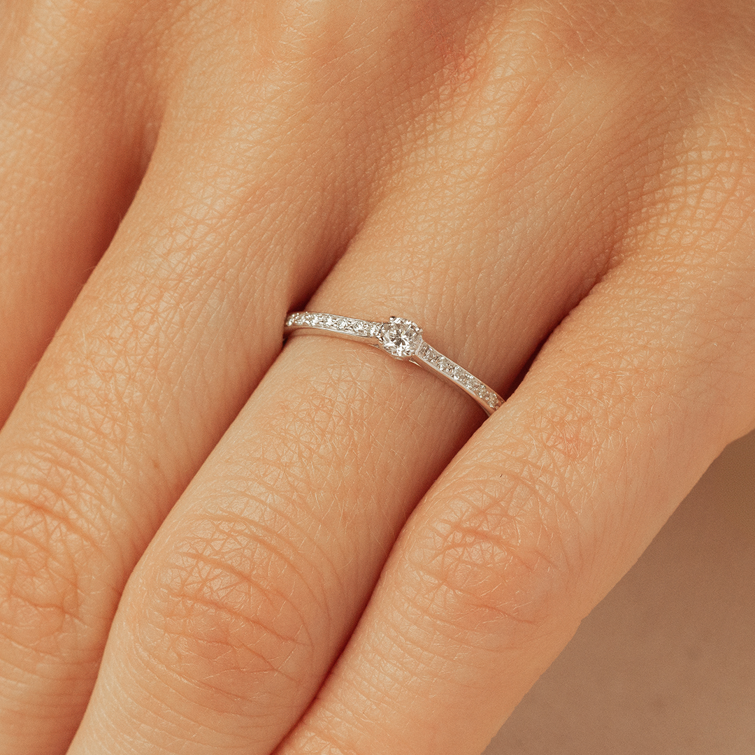 Bague en or blanc ornée d'un diamant central et de petites pierres scintillantes sur le bandeau, portée sur un doigt humain. Ce modèle élégant et moderne met en valeur la beauté du diamant de manière délicate, idéale pour des occasions spéciales telles que des fiançailles ou des célébrations.