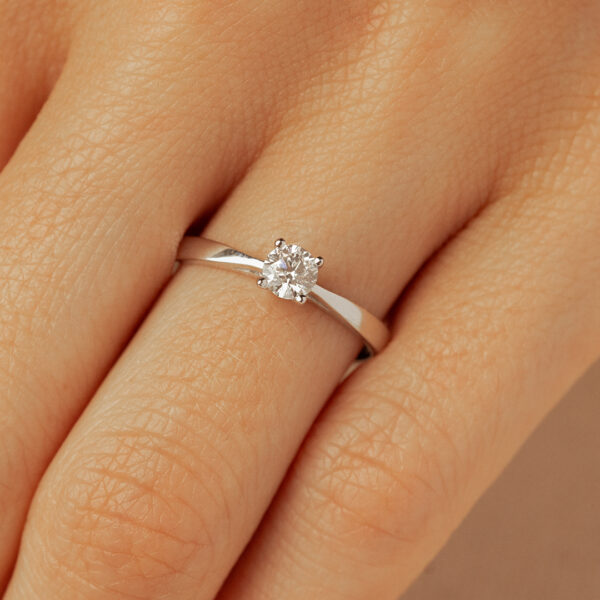 Bague en diamant solitaire délicate, mise en valeur sur un doigt. La bague en or blanc arbore un brillant éclatant, idéal pour une demande en mariage ou un cadeau spécial. Design moderne et élégant, parfait pour toute occasion.