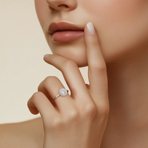 Une magnifique bague en diamant rond, brillamment sertie, portée sur le doigt d'une main délicate. La main repose sur le visage d'une femme aux lèvres douces et à la peau éclatante, soulignant l'élégance de la bague et l'harmonie de l'ensemble. Idéal pour ceux qui cherchent une bague de fiançailles ou un bijou raffiné.