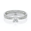 Bague de fiançailles élégante en or blanc avec un diamant central rond brillant entouré de petits diamants sur les côtés, mise en valeur par un design raffiné et moderne, parfaite pour une proposition romantique.