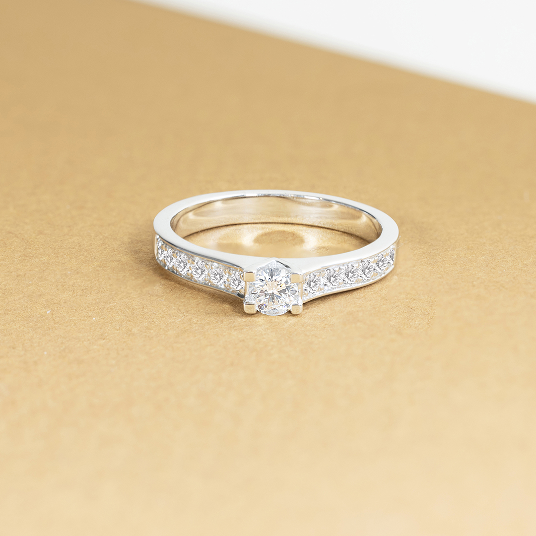 Bague en or blanc sertie d'un diamant central brillant et ornée de pierres précieuses sur les côtés, idéale pour une proposition de mariage ou une occasion spéciale, élégante et sophistiquée avec une touche moderne.