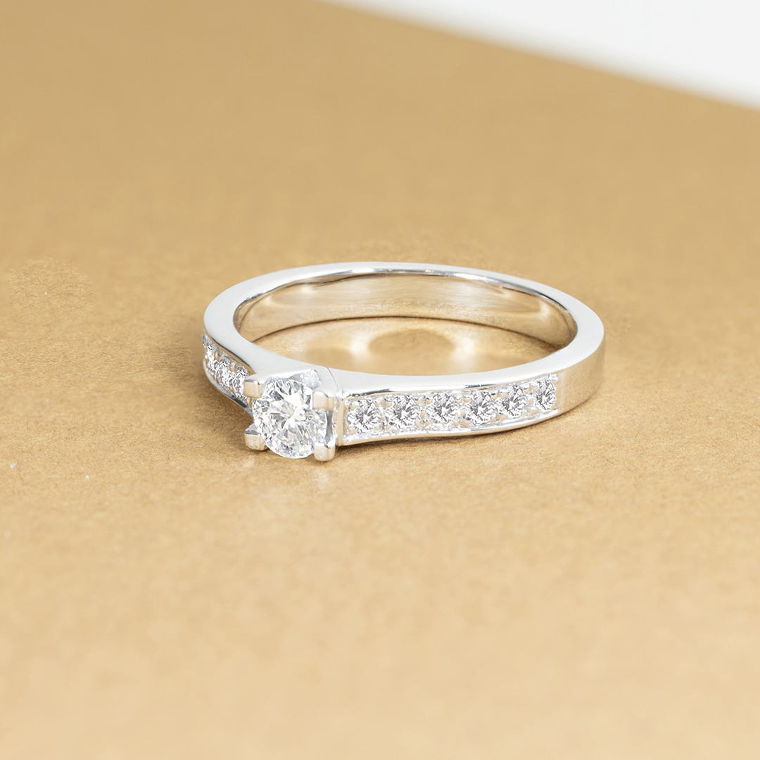 Bague en or blanc avec un diamant central taillé en ancienne, entourée de petits diamants sur le côté. Cette élégante bague de fiançailles brille sur un fond doré, mettant en valeur son design sophistiqué et moderne. Idéale pour un engagement romantique ou une occasion spéciale.