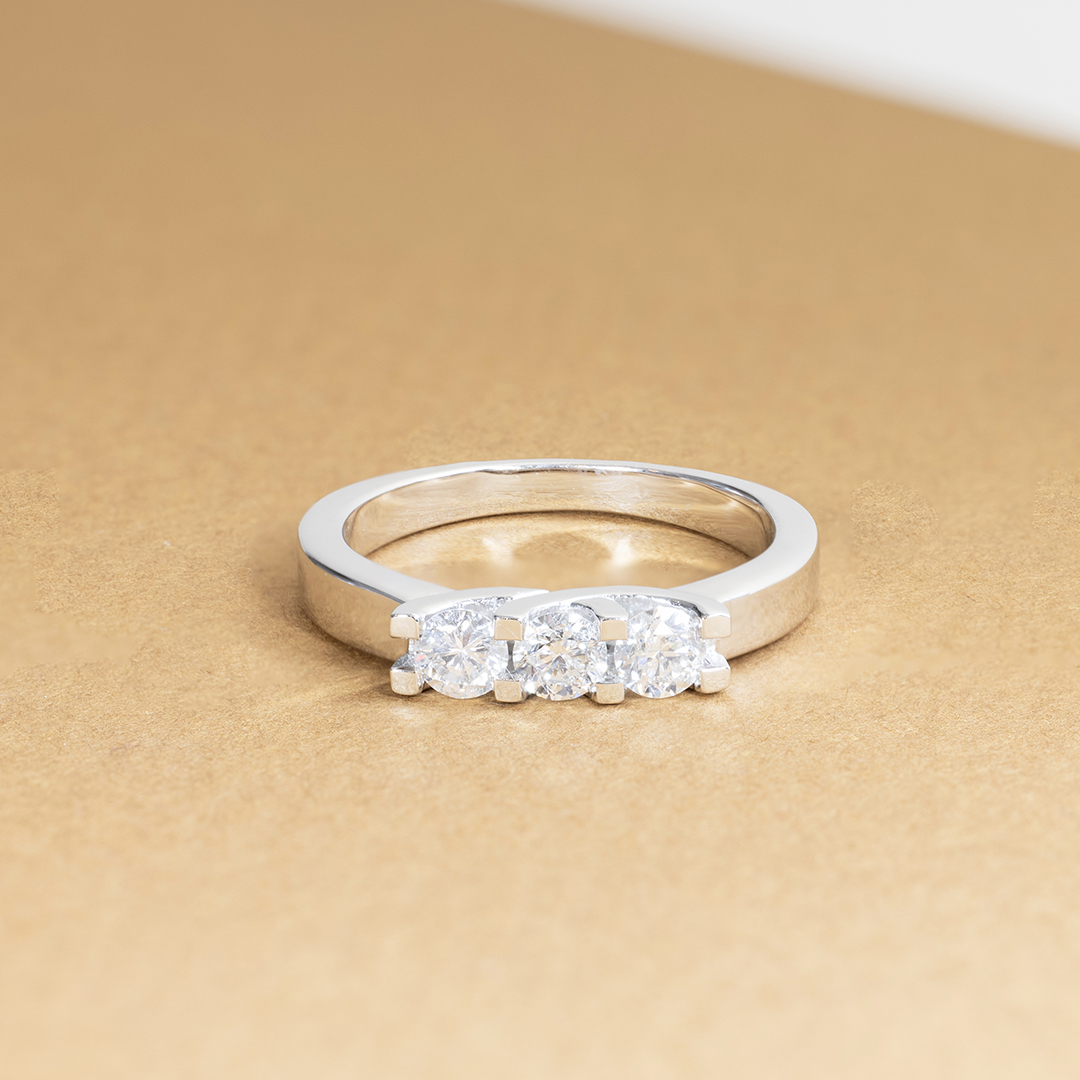Bague en argent avec trois diamants ronds scintillants, montée sur un anneau lisse, présentée sur un fond doré, parfaite pour une bague de fiançailles ou un cadeau spécial, mettant en valeur l'éclat et l'élégance des pierres précieuses.