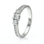 Bague de fiançailles en or blanc avec un diamant central brillant entouré de petits diamants sur les côtés, design élégant et moderne, symbole d'amour éternel, parfaite pour un engagement ou une occasion spéciale.