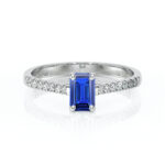 Bague en or blanc sertie d'une pierre précieuse saphir bleu de taille émeraude, entourée de diamants brillants sur chaque côté, parfaite pour une demande en mariage ou une occasion spéciale, modèle élégant et intemporel, mettant en valeur la beauté et l'éclat du saphir.