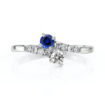 Bague en or blanc ornée d'un saphir bleu rond et d'un diamant central brillant, avec des diamants en ligne sur le corps de la bague, symbole d'élégance et de sophistication. Parfait pour une demande en mariage ou une occasion spéciale, ce modèle de bague allie modernité et classicisme.