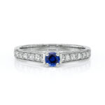 Bague en or blanc ornée d'une pierre précieuse saphir bleu centrale, entourée de diamants scintillants sur le bandeau. Élégante et raffinée, idéale pour les fiançailles ou les occasions spéciales. Design contemporain mettant en avant le contraste entre le bleu du saphir et l'éclat des diamants.