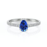 Bague en diamant avec une pierre précieuse en forme de poire, de couleur bleue, sertie en centre, accompagnée de diamants scintillants sur la bande en argent. Cette magnifique bague est idéale pour une demande en mariage ou une occasion spéciale, symbolisant l'élégance et l'amour éternel.