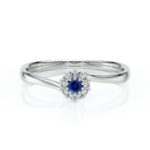 Bague en or blanc avec un motif élégant, ornée d'un saphir bleu central entouré de diamants scintillants, parfaite pour un engagement ou une occasion spéciale. Modèle romantique avec une finition brillante, cette bague évoque le luxe et la sophistication.