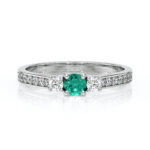 Bague en or blanc avec une pierre précieuse centrale en émeraude, entourée de deux diamants, ornée d'une bande de diamants sur le côté, idéale pour une occasion spéciale ou comme bague de fiançailles, mettant en avant le contraste entre l'émeraude verte lumineuse et les diamants scintillants, ajoutant une touche d'élégance et de sophistication.