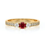 Bague en or jaune ornée d'un rubis central entouré de deux diamants ronds, avec un pavé de diamants sur l'anneau, symbole d'élégance et de luxe, idéal pour une déclaration d'amour ou un cadeau spécial.